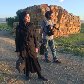 Sara Khaki and Mohammad Reza Eyni walking on a road next to piles of straw. Sara has a camera hanging from her neck and Mohammad audio recording equipment.