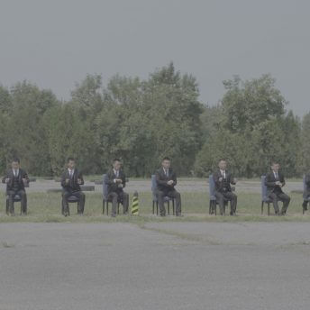 Still from Ascension. Full shot of 11 men training to become body guards. They are sitting on chairs in an open field, wearing suits.