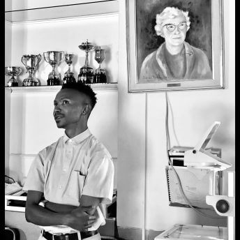 Still from Milisuthando (working title). A man is standing in front of a bookshelf with crossed arms. There is a portrait of an elderly woman to his left and trophies on a shelf to his right. Black and white photo.