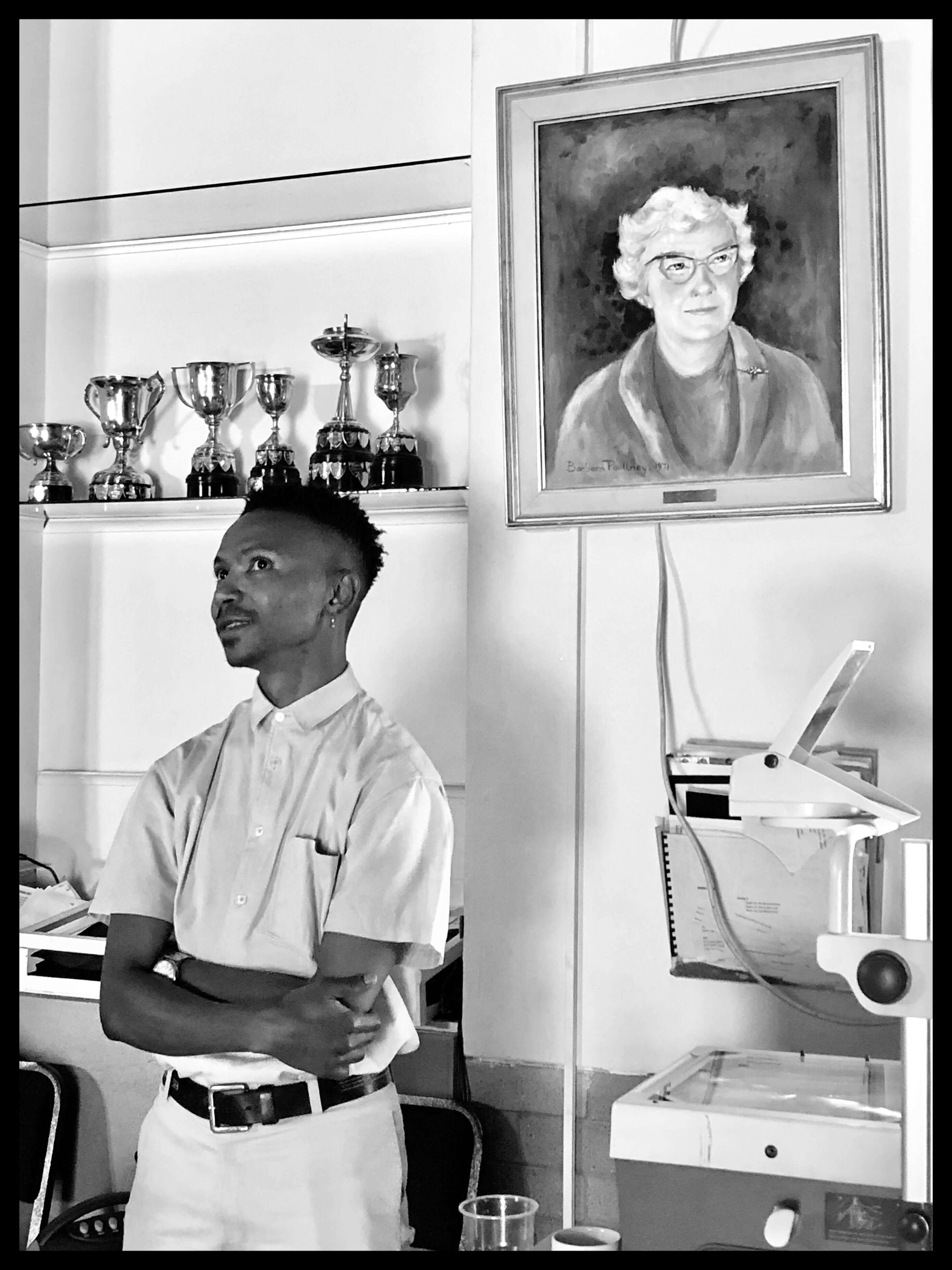 Still from Milisuthando (working title). A man is standing in front of a bookshelf with crossed arms. There is a portrait of an elderly woman to his left and trophies on a shelf to his right. Black and white photo.