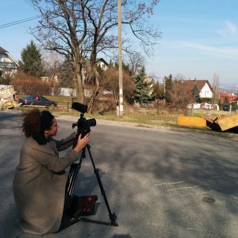 Farah Kassem kneels on a suburban street positioning a camera on a tripod. It's a sunny Autumn day. Color photograph.
