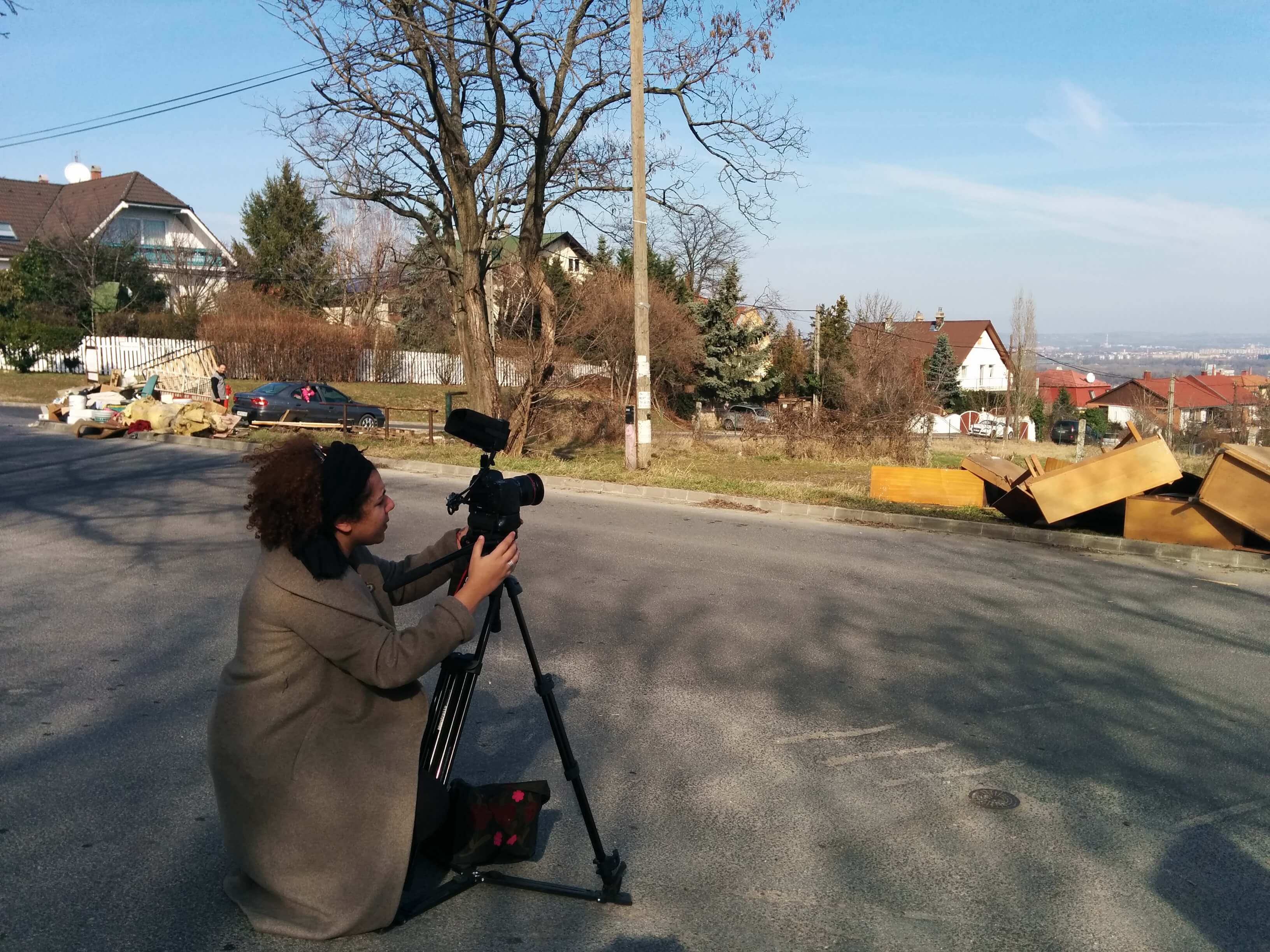 Farah Kassem kneels on a suburban street positioning a camera on a tripod. It's a sunny Autumn day. Color photograph.
