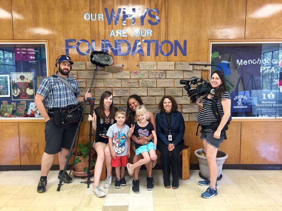 Production still from Mama Bears. Seven people are in front of a wall that says, "Our Whys Are Our Foundation". There are three crew members with camera equipment. Two crew members are standing and one is sitting on a bench with two women and two children.