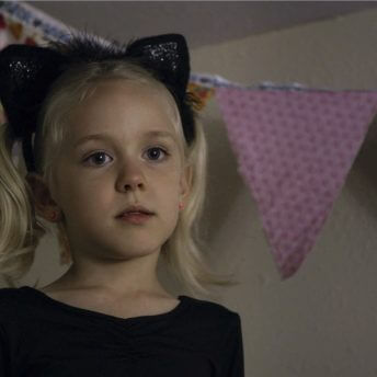 Still from Mama Bears. A young, blonde girl with a headband of black cat ears stands in front of a hanging banner of various patterned fabric flags.