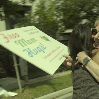 Still from Mama Bears. Two women are hugging. One woman, wearing sunglasses, is holding a sign that reads, "Free Mom Hugs" as well as more text that is not legible.