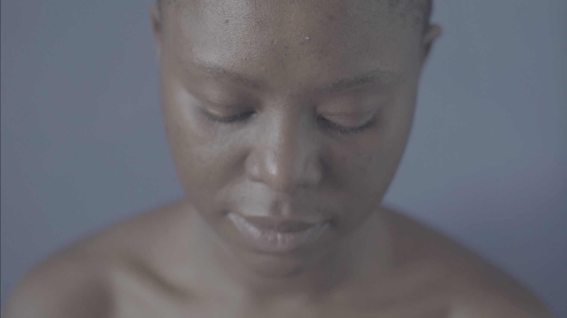 Still from Milisuthando (working title). A close-up, downward angle of a woman who is looking down and away from the camera. The background is blue.