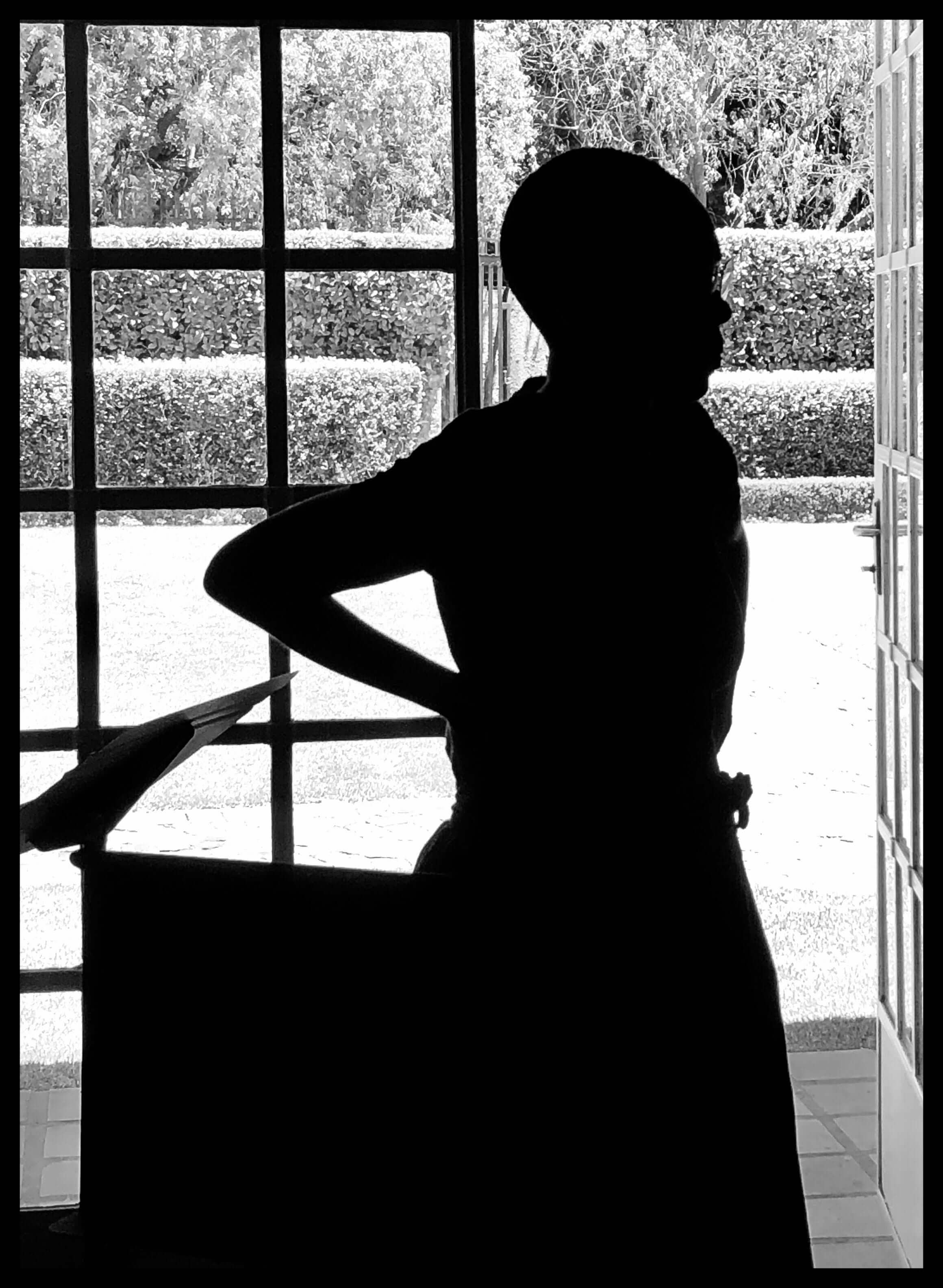 Still from Milisuthando (working title). A person is standing with their hands on their hips in front of a windowed door open to an outside garden area. Backlit, black and white photo.