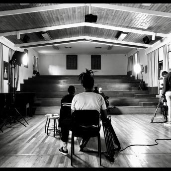 Production still from Milisuthando (working title). Two crew members are sitting infront of and filiming Milisuthando Bongela. Another camera man is filming from a different angle on the right. A few lights are surrounding the participant.