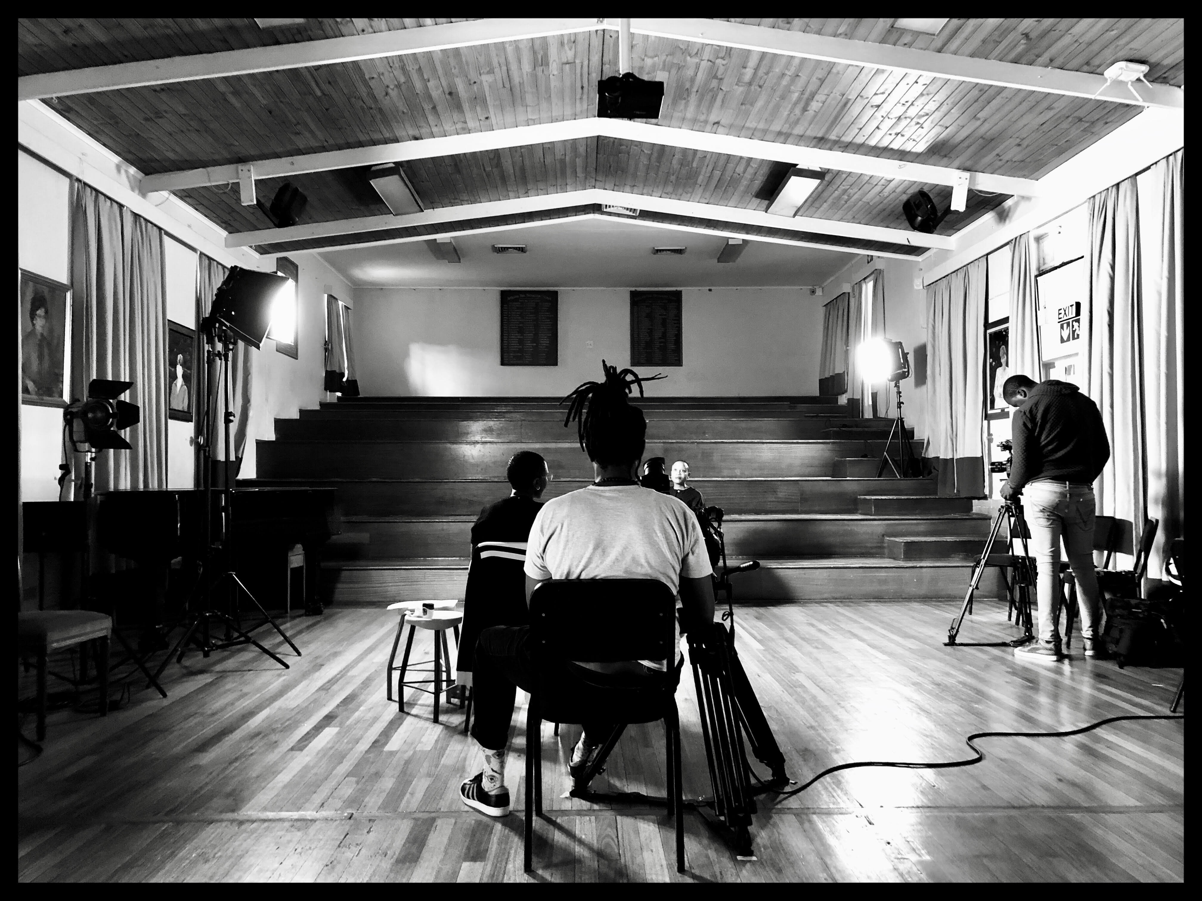 Production still from Milisuthando (working title). Two crew members are sitting infront of and filiming Milisuthando Bongela. Another camera man is filming from a different angle on the right. A few lights are surrounding the participant.