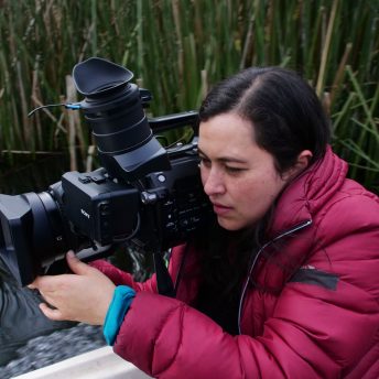 Production still of Paths of Fire. Filmmaker Viviana Gómez Echeverry works with a camera, she is wearing a red puffed jacket.