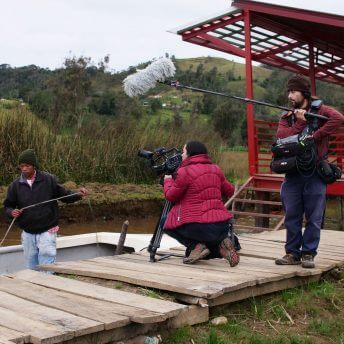 Production still of Paths of Fire. Filmmaker Viviana Gómez Echeverry is working with a camera and with a sound crew person. They are filming a man working on a boat.