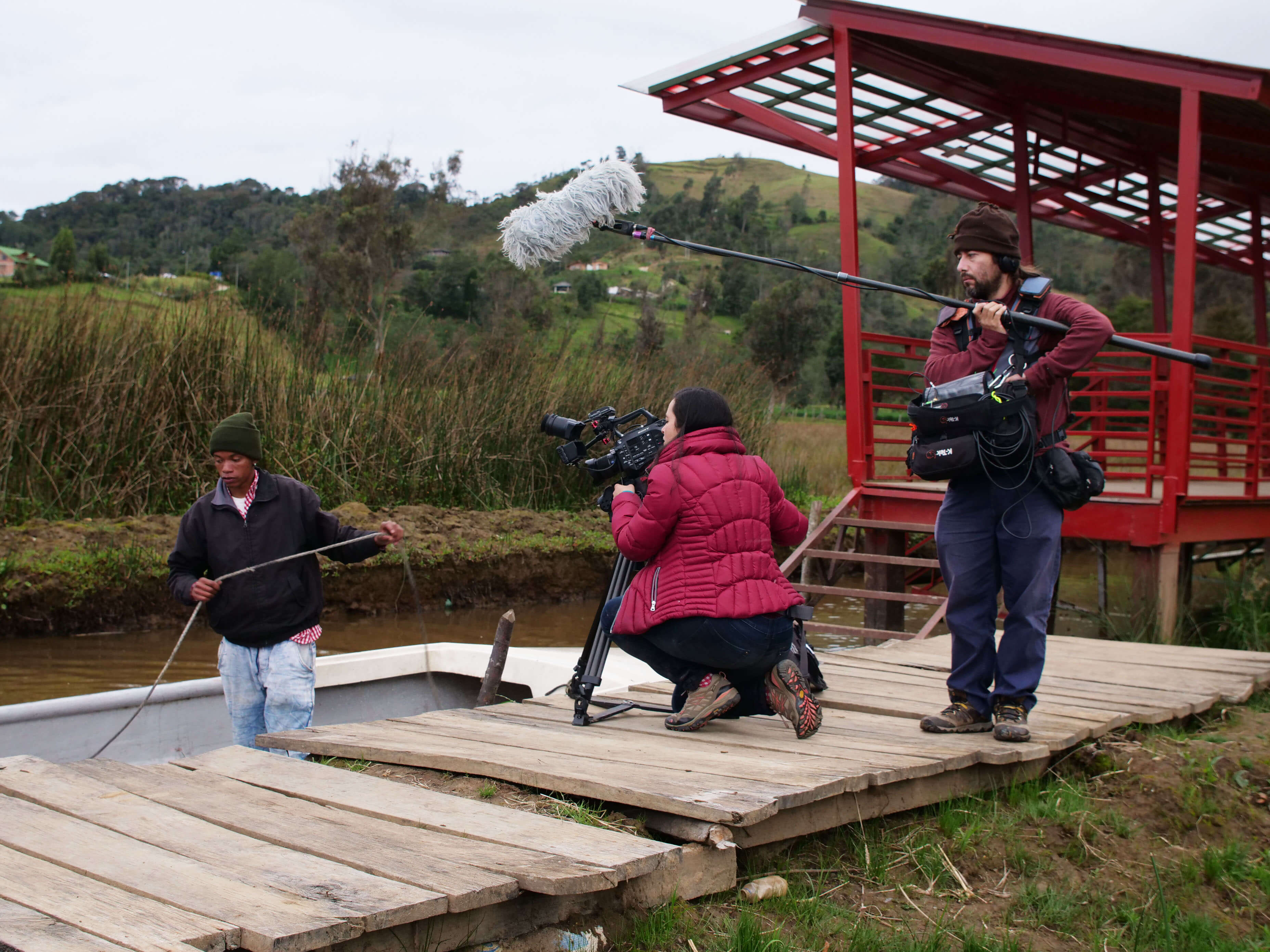 Production still of Paths of Fire. Filmmaker Viviana Gómez Echeverry is working with a camera and with a sound crew person. They are filming a man working on a boat.