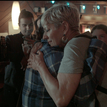 Still from Mama Bears. Two women are hugging. One woman, wearing sunglasses, is holding a sign that reads, "Free Mom Hugs" as well as more text that is not legible.