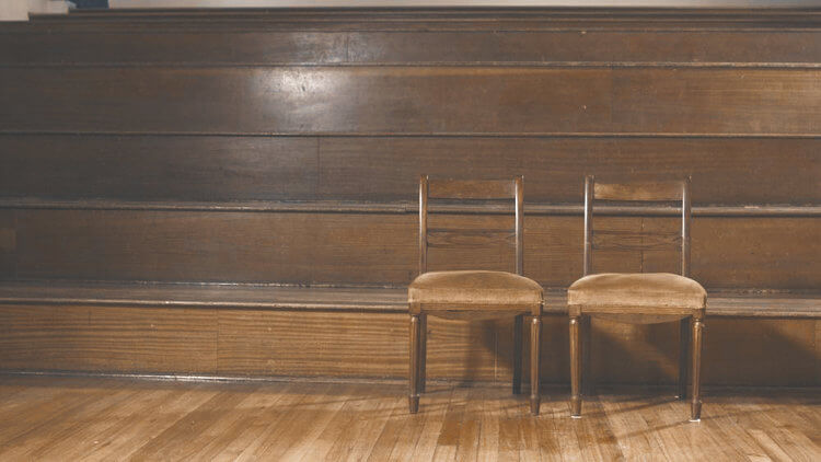 Still from Milisuthando (working title). Two wooden chairs sit infront of wooden stadium style bench seating.