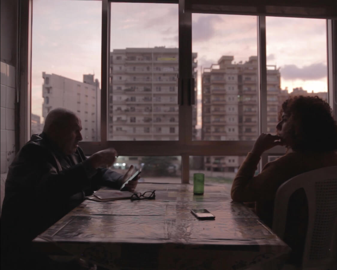 Farah Kassem and her father sit across from each other at a table. Farah's father Mustapha is looking at his phone. In the background, tall buildings can be seen through the large window. Color photograph.