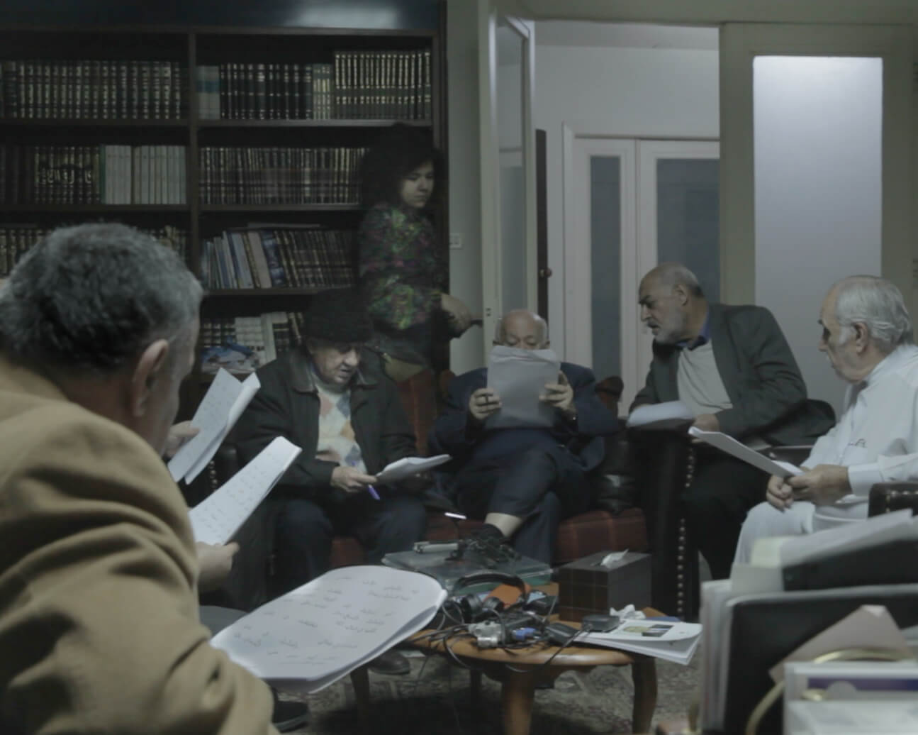 A group of men sits on couches holding papers in front of them. Farah Kassem is walking in the background next to a bookshelf. Color photograph.