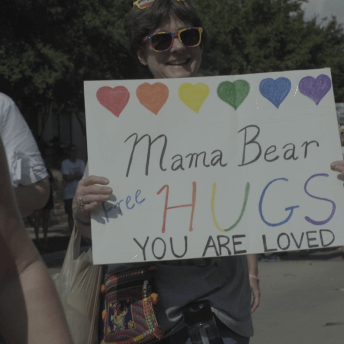 Still from Mama Bears. A woman in sun glasses holds a sign with hearts in rainbow colors that reads, "Mama Bear Free Hugs. You are Loved". There is a crowd of people around and behind her.