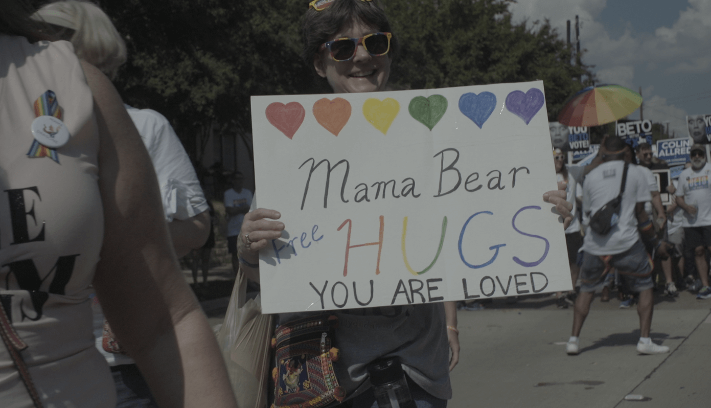 Still from Mama Bears. A woman in sun glasses holds a sign with hearts in rainbow colors that reads, "Mama Bear Free Hugs. You are Loved". There is a crowd of people around and behind her.