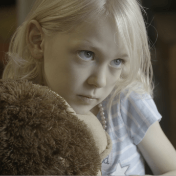 Still from Mama Bears. A young, blonde girl is holding a large stuffed animal and looking off right of the camera.