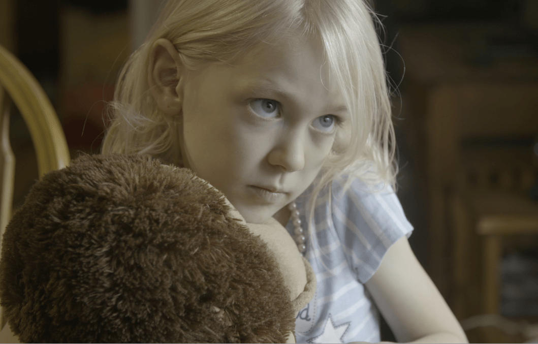 Still from Mama Bears. A young, blonde girl is holding a large stuffed animal and looking off right of the camera.