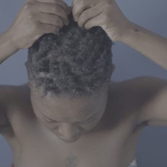 Still from Milisuthando (working title). A camera angle pointing down shows a woman who is touching the top of her head. The background is blue.