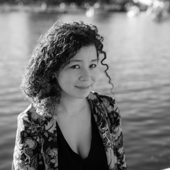 Jessica Kingdon is in front of a lake. She is gently smiling. Her hair is curly, and she wears a blazer and a top. Portrait in black and white.