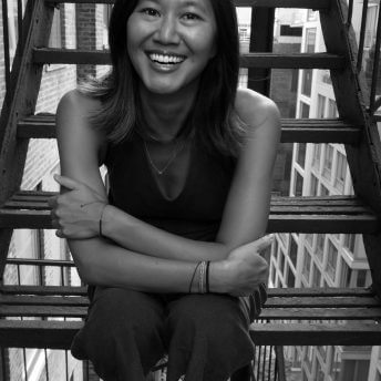 Stephanie Wang-Breal sitting on fire escape stairs, smiling widely. She has mid-shoulder length, dark hair, and wears a sleeveless shirt. Black and white portrait.