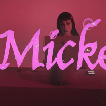 Still form #Mickey. A woman sits behind a desk with a computer screen and cell phone on it. The tile of the film, "#Mickey" overlays the image in pink with sparkles.