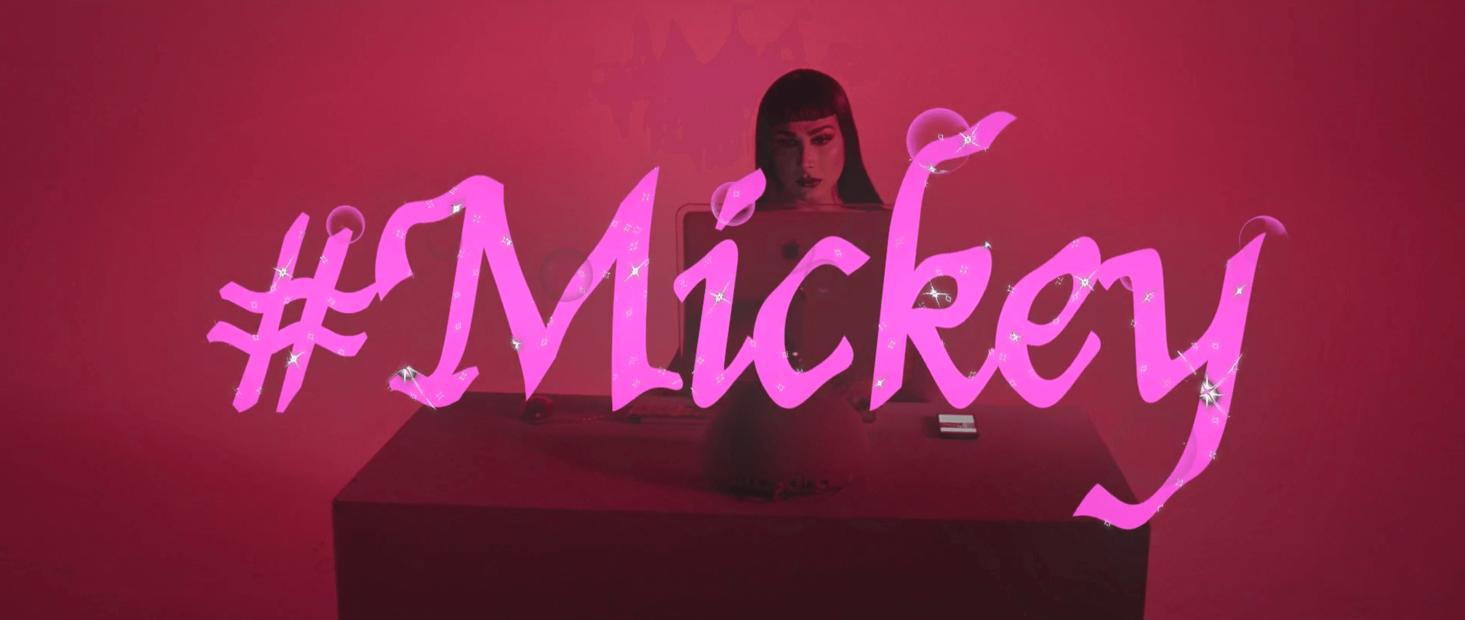 Still form #Mickey. A woman sits behind a desk with a computer screen and cell phone on it. The tile of the film, "#Mickey" overlays the image in pink with sparkles.