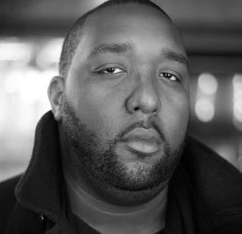 Jason Pollard looking at the camera, wearing a dark jacket with the collar upturned. Black and white portrait, with the background of out of focus.