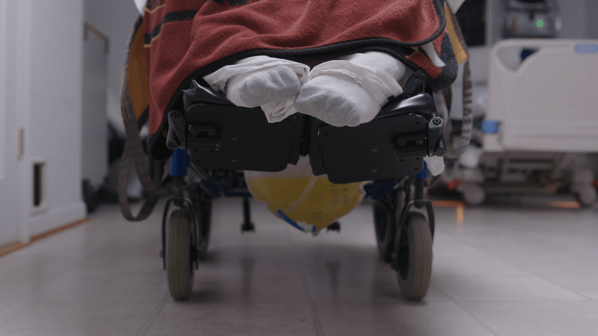 A photo of a person sitting in a wheelchair, only their feet are visible and they are wearing white socks. A red blanket over their lap.