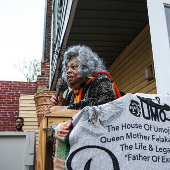 A woman with grey curly hair and a breathing tube connected to her nose is holding a microhpone. In the foreground hangs a grey fabric sign with black text that reads "The House of Umoja Along with Queen Mother Falaka Fattah Honors the Life & Legacy of Our "Father of Excellence"."