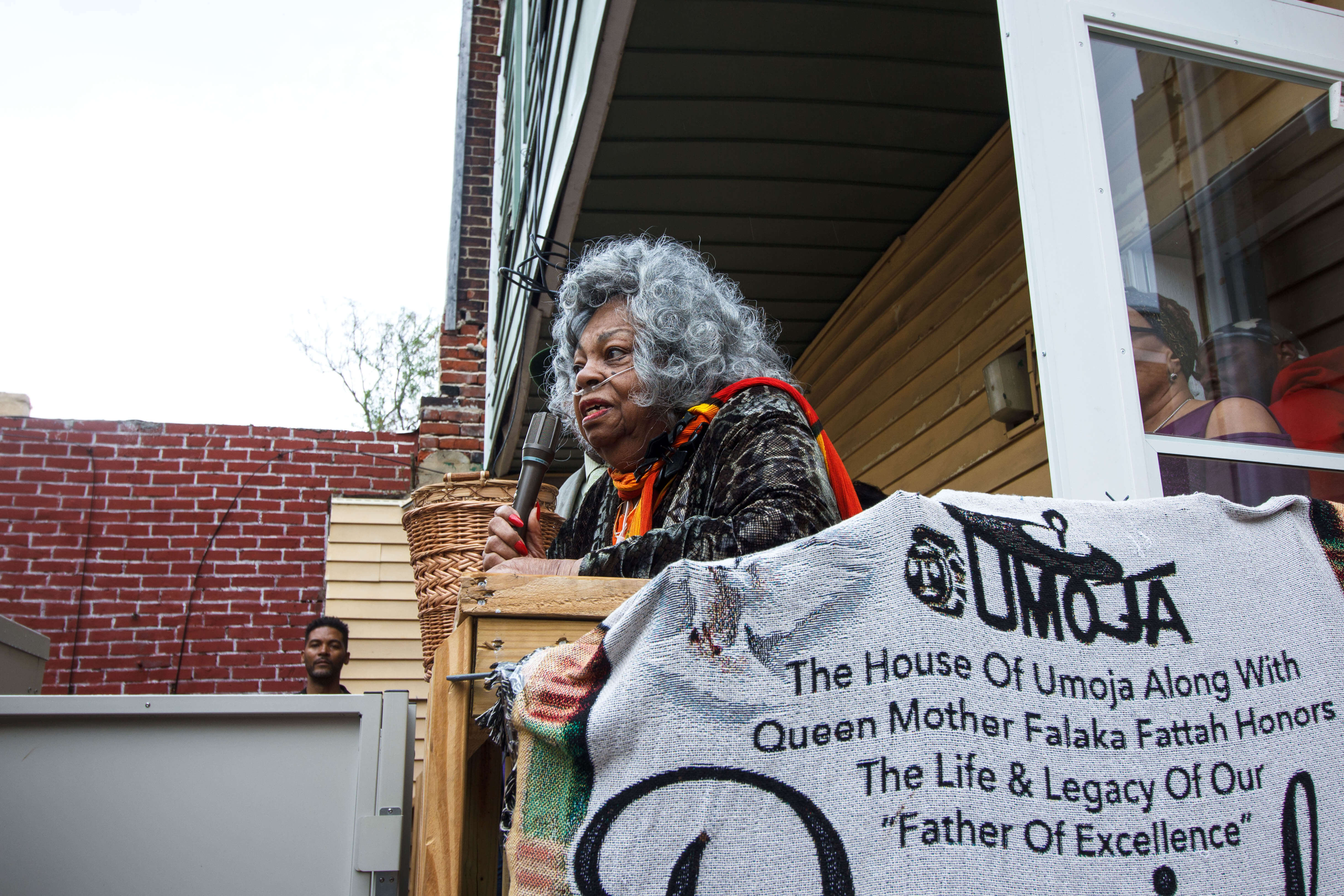 A woman with grey curly hair and a breathing tube connected to her nose is holding a microhpone. In the foreground hangs a grey fabric sign with black text that reads "The House of Umoja Along with Queen Mother Falaka Fattah Honors the Life & Legacy of Our "Father of Excellence"."