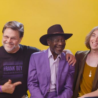 Production still from Frank Bey: You're Going to Miss Me. Frank Bey–a man in a wide-brimmed brown hat and purple pantsuit-- is seated between two other people. The person to the right of him wears a t-shirt that has the text "Frank Bey: Back in Business" on it, and has one arm around the shoulder of the man in the middle, and with the other arm is pointing towards the man in the middle. The person to the left of him is the director, Marie Hinson. Partial view of a living space is seen on either edge of the golden-yellow backdrop.