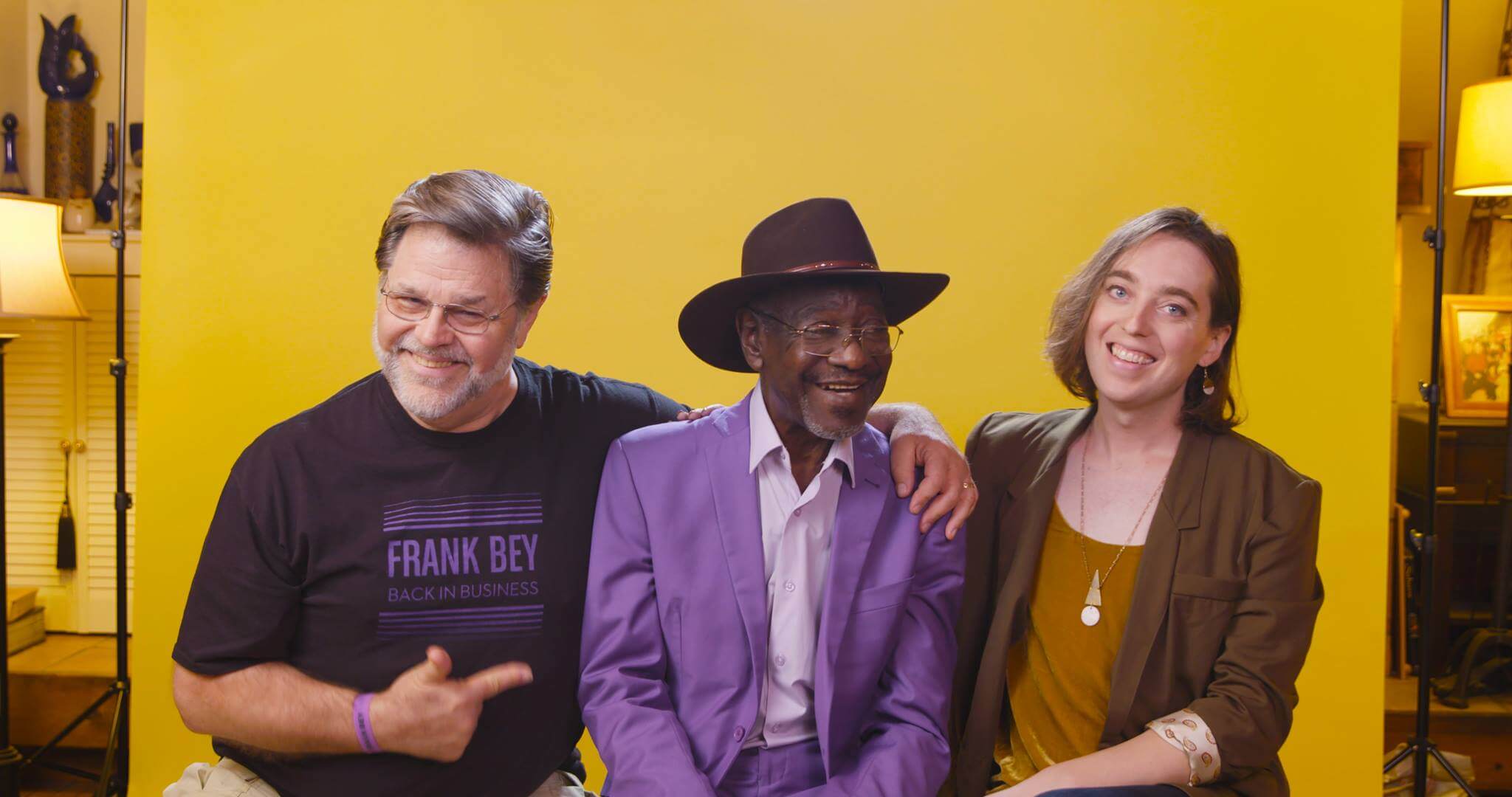 Production still from Frank Bey: You're Going to Miss Me. Frank Bey–a man in a wide-brimmed brown hat and purple pantsuit-- is seated between two other people. The person to the right of him wears a t-shirt that has the text "Frank Bey: Back in Business" on it, and has one arm around the shoulder of the man in the middle, and with the other arm is pointing towards the man in the middle. The person to the left of him is the director, Marie Hinson. Partial view of a living space is seen on either edge of the golden-yellow backdrop.