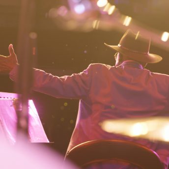 Still from Frank Bey: You're Going to Miss Me. Seen from behind: Frank Bey–a man in a wide brimmed hat and jacket with his arms stretched wide. To his left is a lectern with papers, and he is illuminaed from the front by bright lights. Parts of a drum set, including a cymbal, are in soft focus in the foreground.