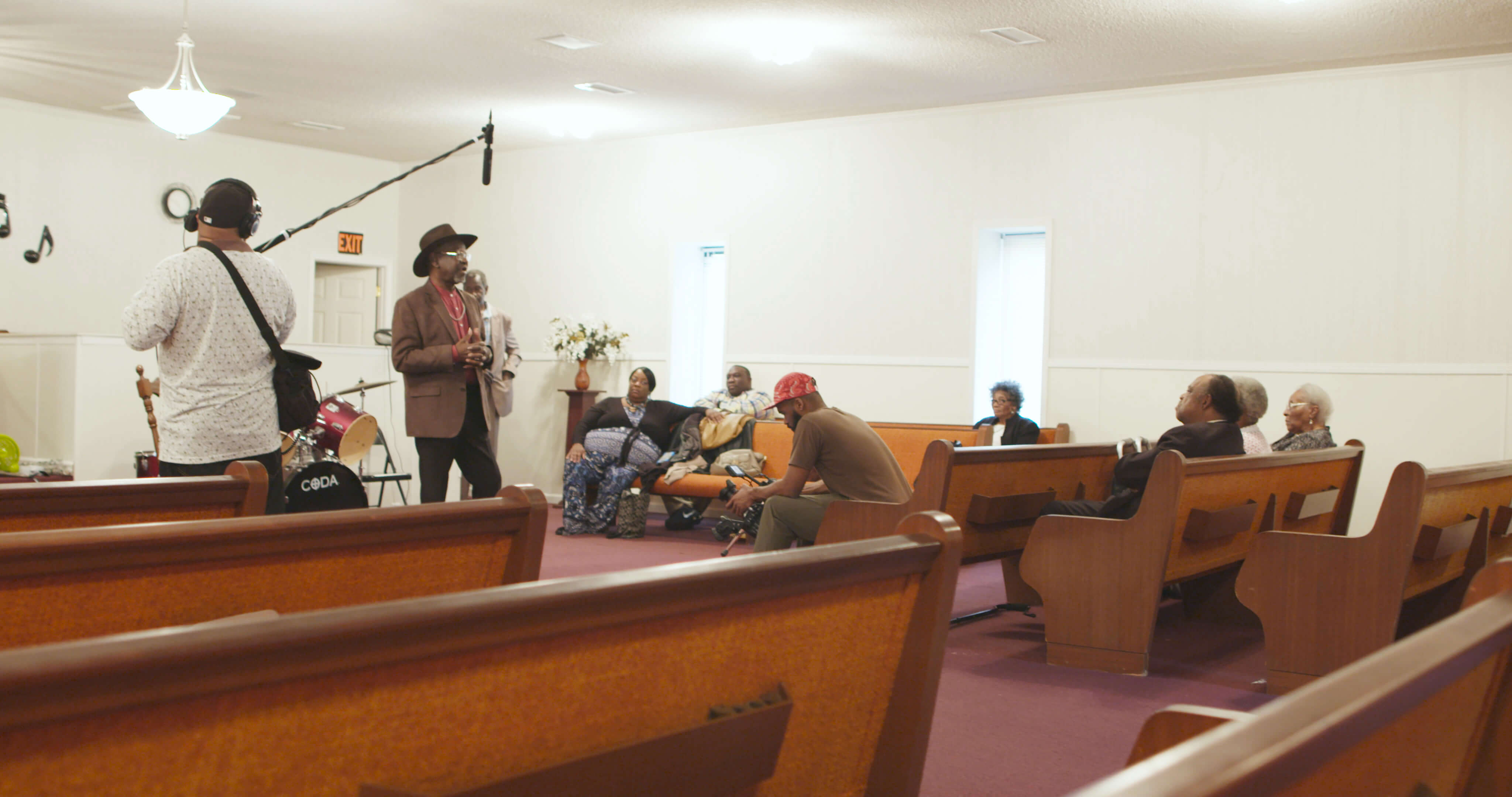 Production still from Frank Bey: You're Going to Miss Me. A man in a brown jacket and wide brimmed hat is addressing a group of people sitting in a wooden pews inside a building. Another is holding a boom mic above him.