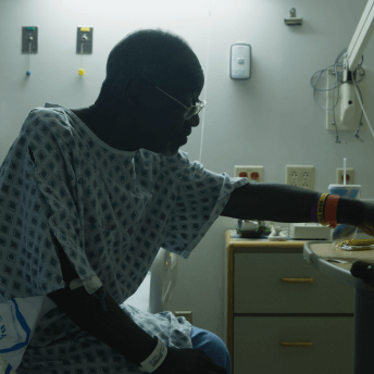 Still from Frank Bey: You're Going to Miss Me. Frank Bey is sitting on a hospital bed wearing a hospital gown. He is reaching with one hand to salties that are on a side table. A wooden cane is also resting on the table.