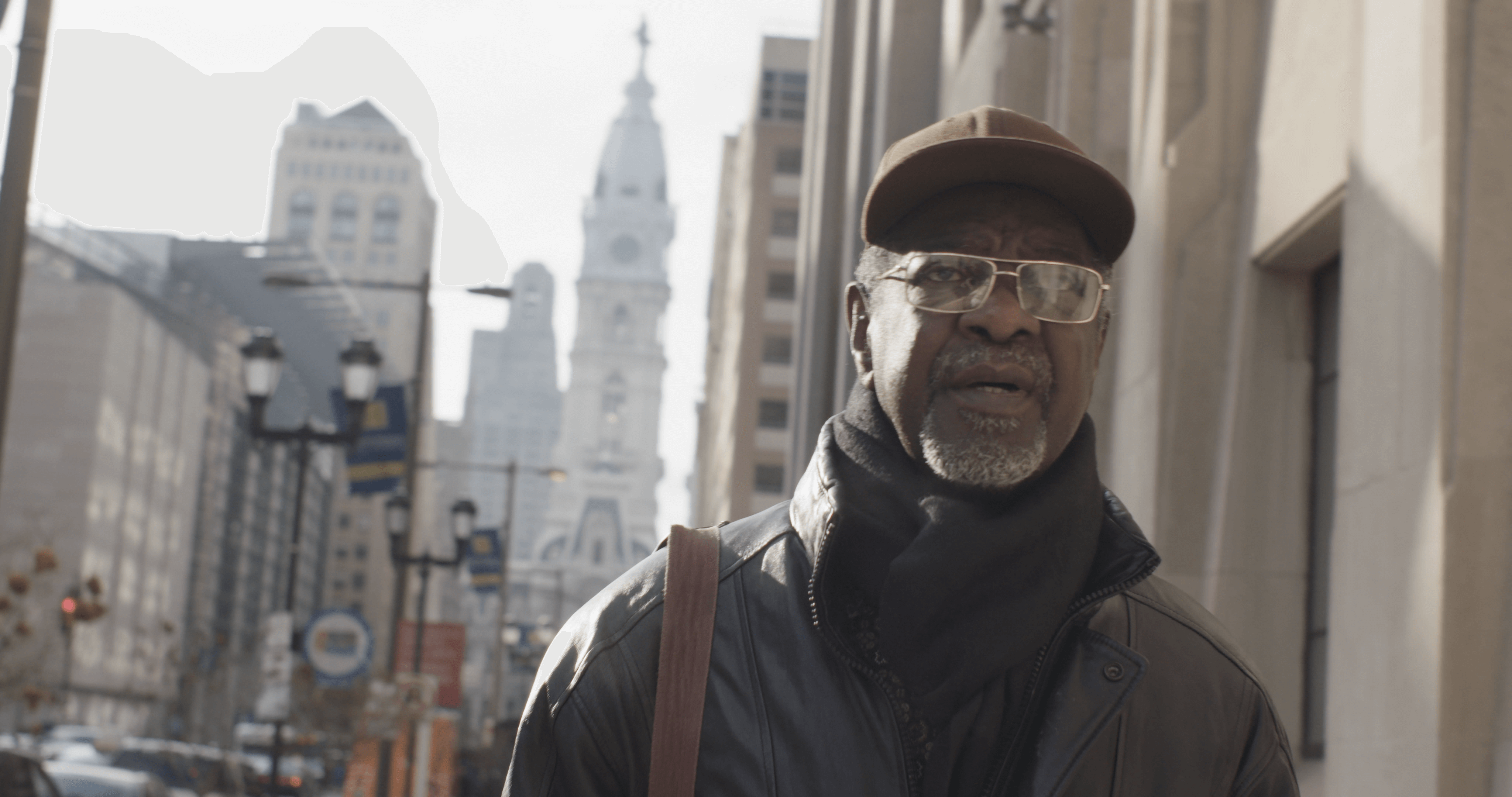 Still from Frank Bey: You're Going to Miss Me. Frank Bey, a man with a salt-and-pepper beard, is wearing gold-frame glasses, brown baseball cap, and black scarf and leather jacket. Behind him tall buildings, street lights, and stoplights are seen slightly out of focus.