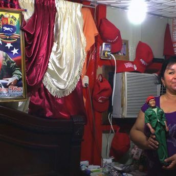Still from Once Upon a Time in Venezuela. A woman stands in the corner of a room by a bed and window air conditioner unit. She is surrounded by political pictures of and merchandise, holding a political figurine of Cesar Chavez.