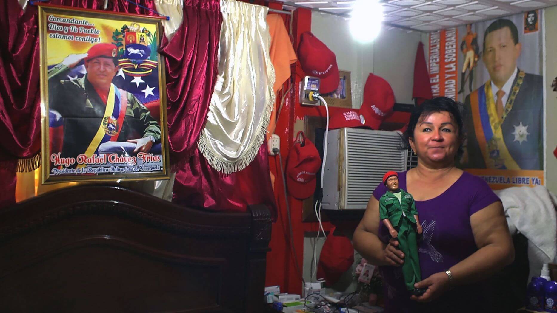 Still from Once Upon a Time in Venezuela. A woman stands in the corner of a room by a bed and window air conditioner unit. She is surrounded by political pictures of and merchandise, holding a political figurine of Cesar Chavez.
