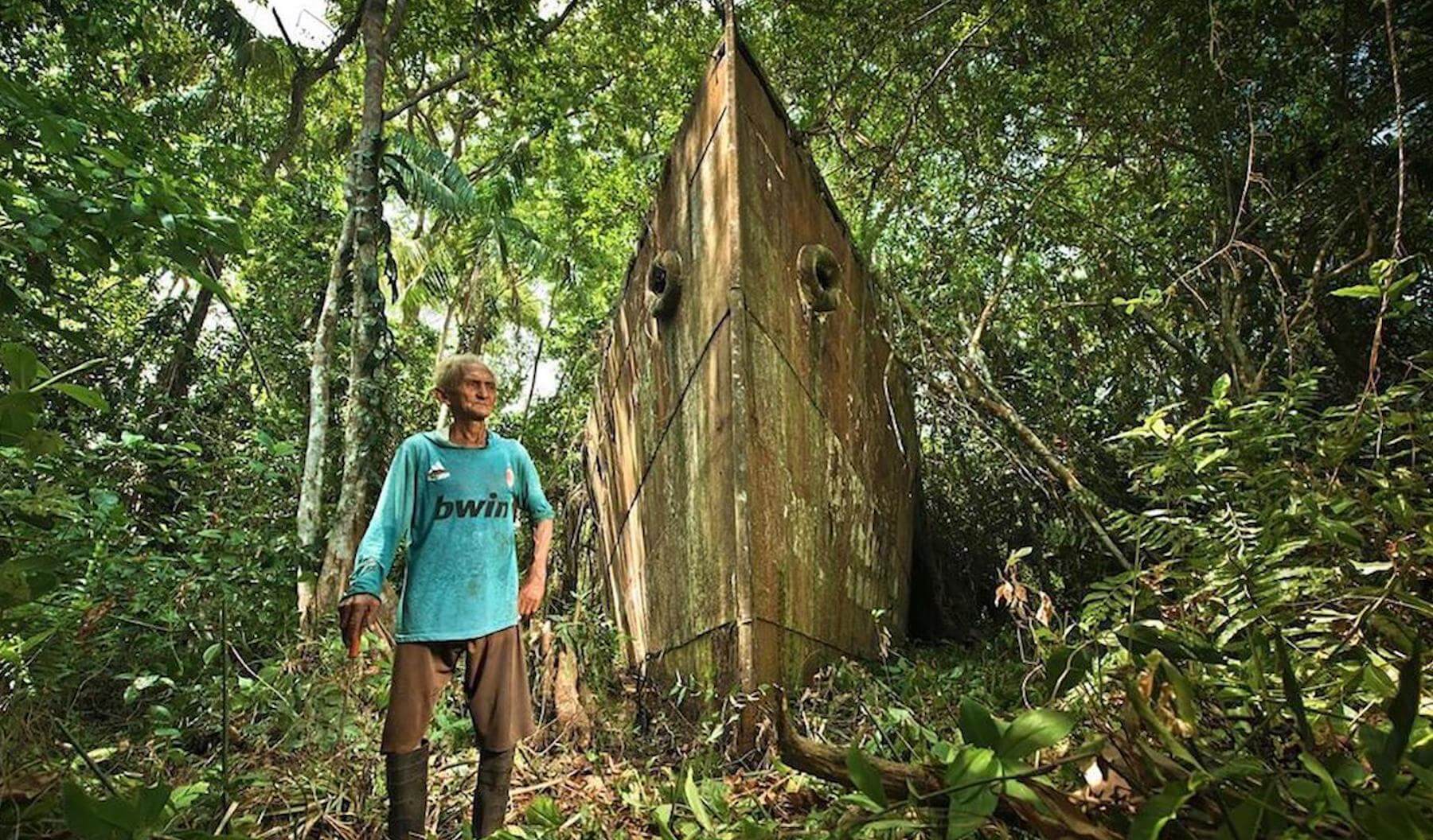 Still from Once Upon a Time in Venezuela. A man stands outside in a forest with an old, rusty boat behind him.