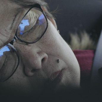 Still from Eskape. A close up of a woman sitting in a car with the seatbelt fastened. She wears glasses, and she is leaning her head to the side with her eyes closed.