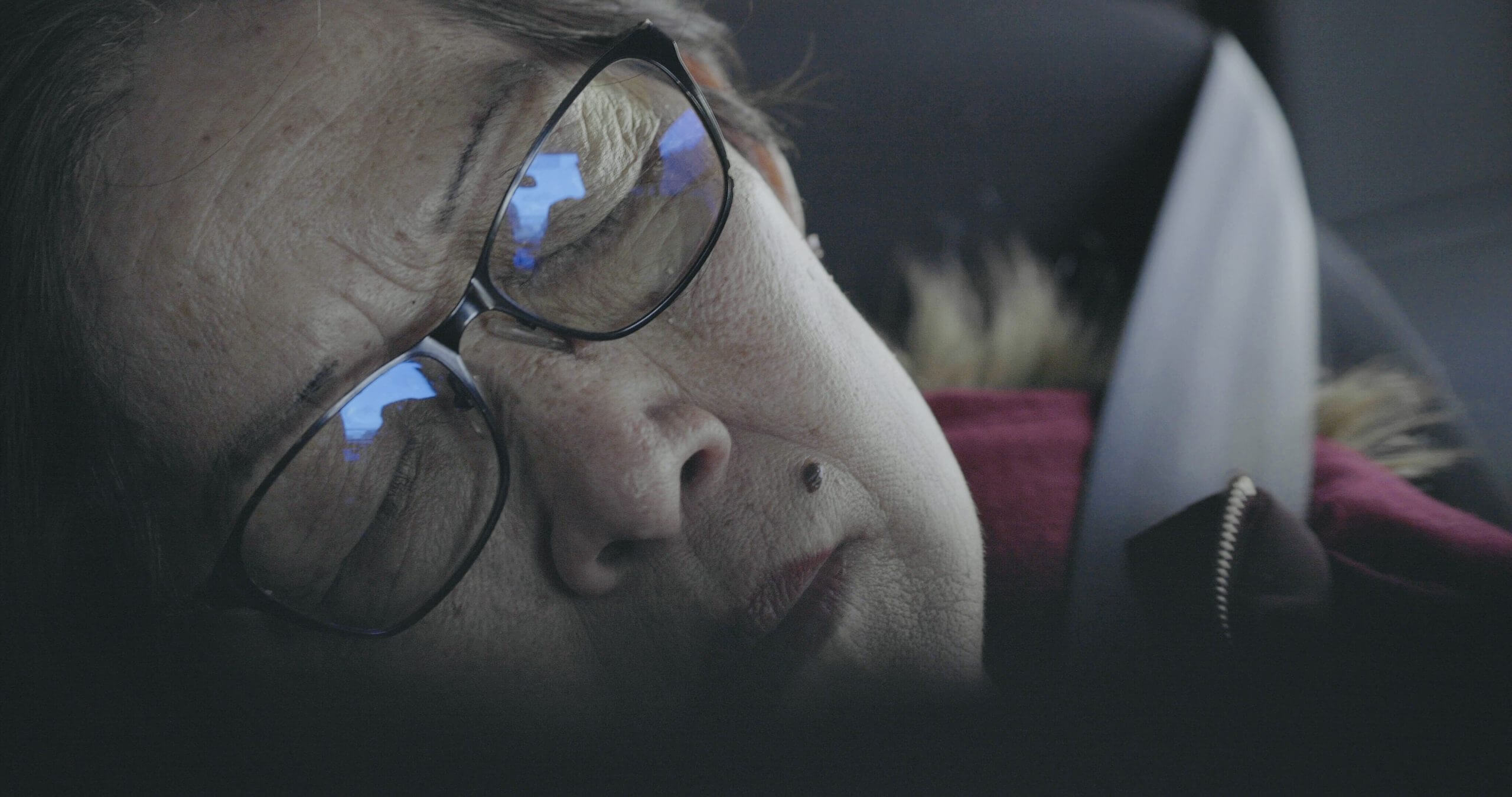 Still from Eskape. A close up of a woman sitting in a car with the seatbelt fastened. She wears glasses, and she is leaning her head to the side with her eyes closed.