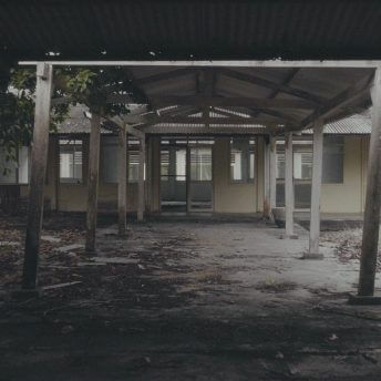 Still from Eskape. Wide shot of the covered entryway to a one-story empty building. The wooden beams supporting the tin metal roof are slightly leaning. The doors and windows of the building are open and the cement walkway in front of the building is partially covered in dirt and moss.