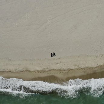 Still from Eskape. Overhead bird-eye view of a beach with waves and water along the bottom one-third. Two dark specks of people are lying on the beach, in the middle of the frame.