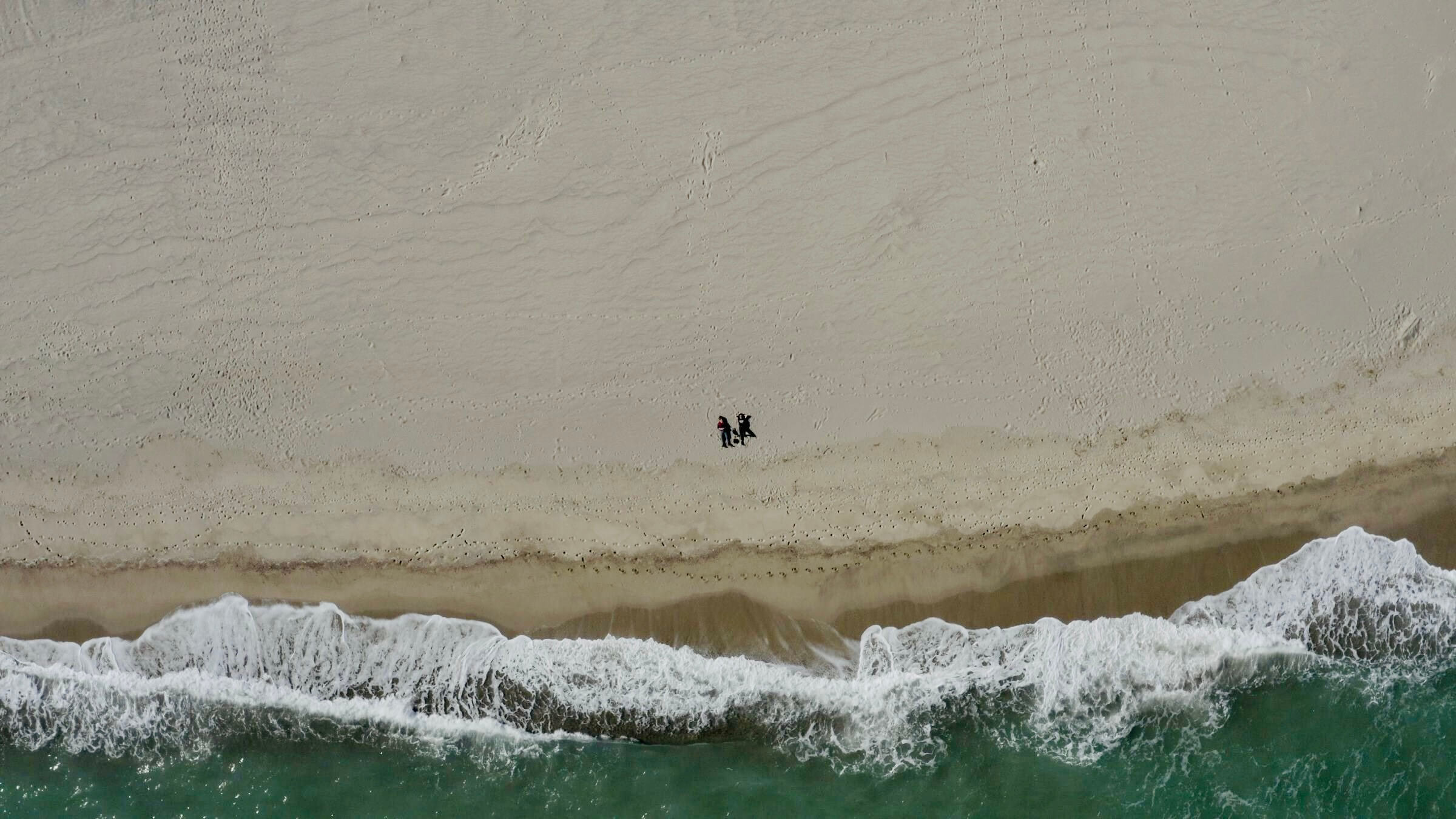 Still from Eskape. Overhead bird-eye view of a beach with waves and water along the bottom one-third. Two dark specks of people are lying on the beach, in the middle of the frame.