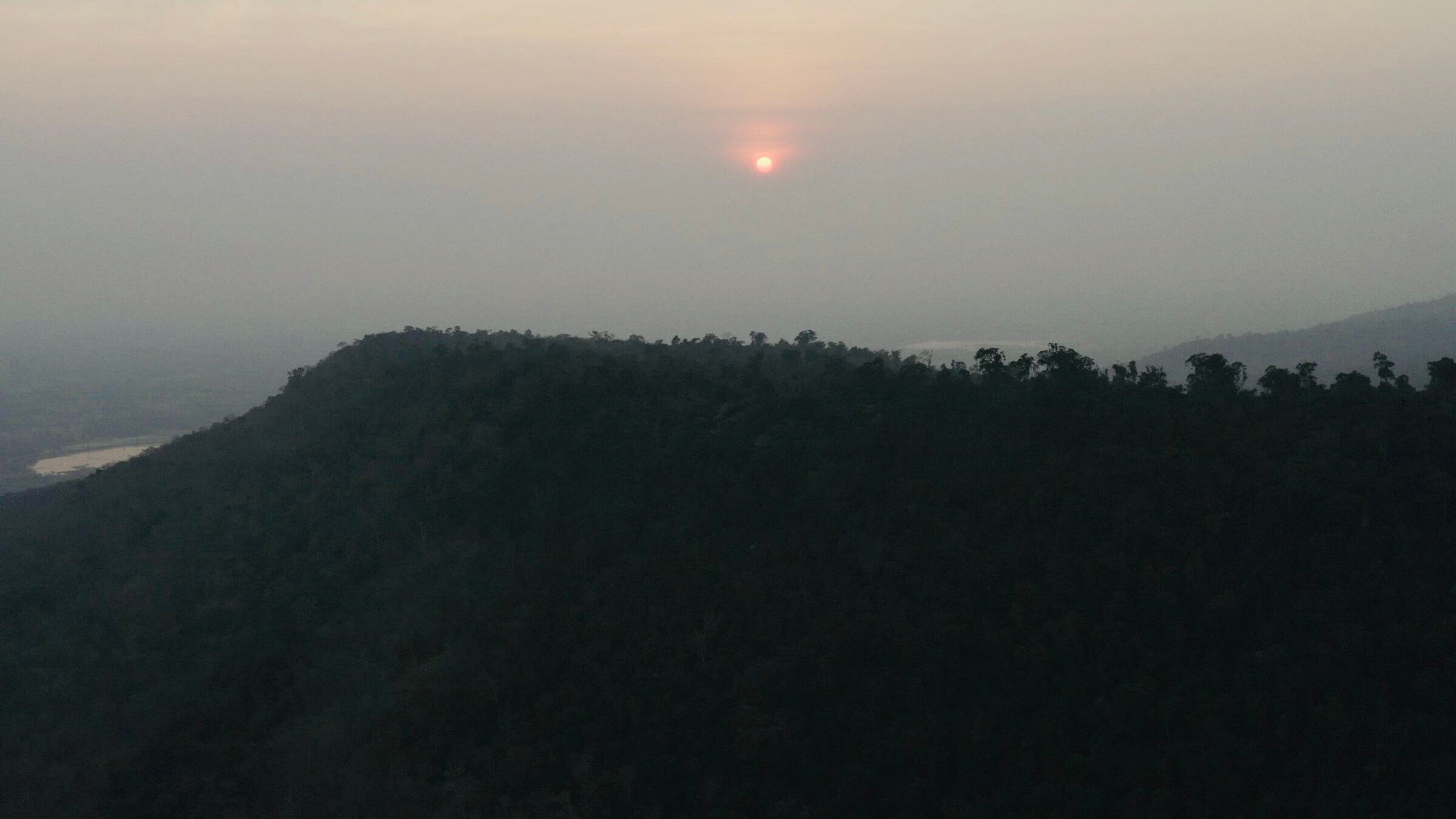 Still from Eskape. A red-orange sun shines over a forest-covered hill, it's rays diffusing out due to the foggy sky.