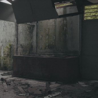 Still from Eskape. Dimly lit, derelict room. Light filters in through the partially-caved in ceiling. There is mildew on the concrete walls and debris and dirt on the floor.