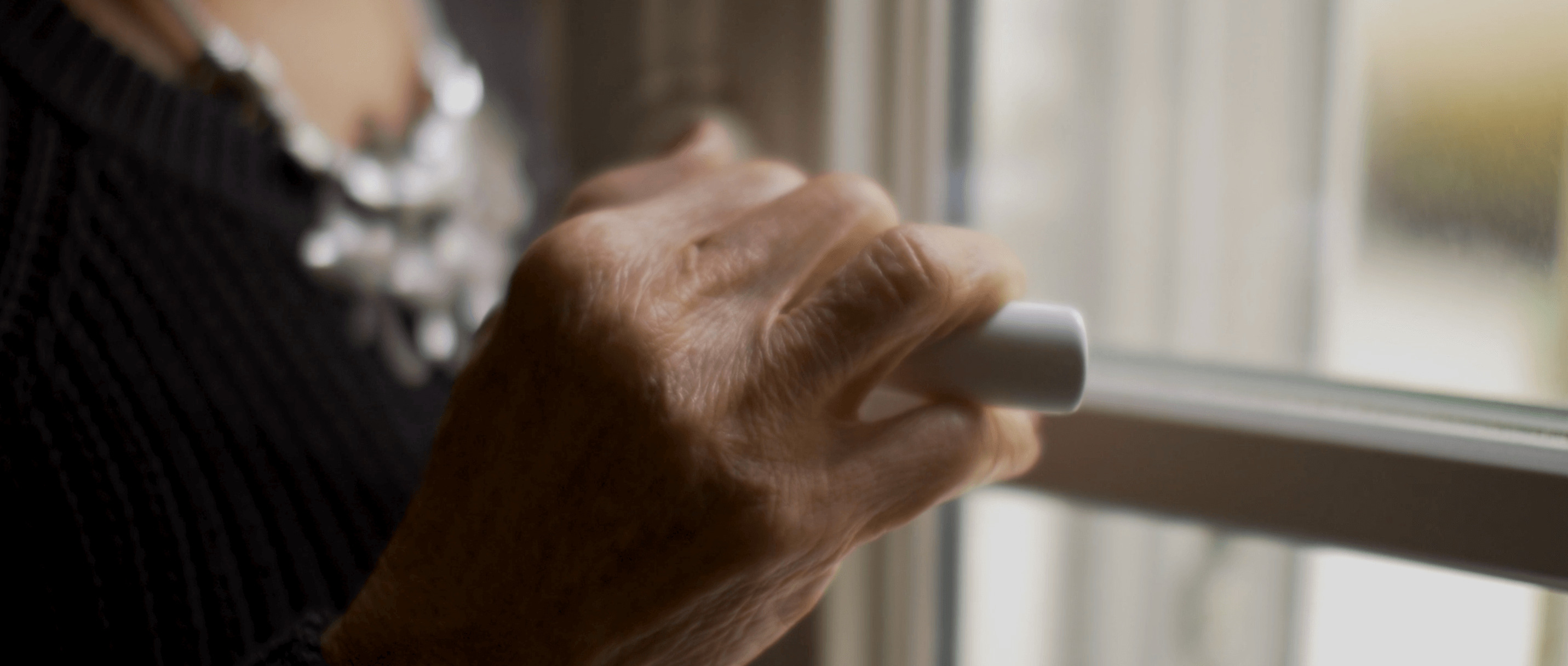 Still from Eskape. Closeup of a woman's hand, holding a window handle as if about to open it. Her hand is in focus, and wrinkles are visible, and her fingernails are painted red. Out of focus, her torso is seen. She is wearing a black sweater and a large silver necklace.